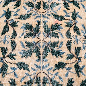 May include: Close-up of a beige rug with a repeating floral pattern. The design features dark green and light blue leaves and vines against a neutral background. The rug has a vintage, slightly worn appearance.