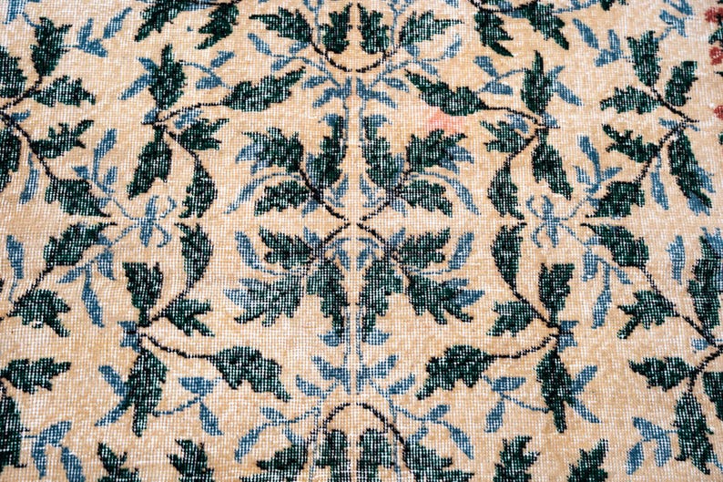 May include: Close-up of a beige rug with a repeating floral pattern. The design features dark green and light blue leaves and vines against a neutral background. The rug has a vintage, slightly worn appearance.