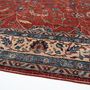 May include: Close-up of a distressed, vintage rug with a rich, reddish-brown field and intricate floral patterns in blue, beige, and other colors. The rug features a detailed border with geometric and floral motifs.