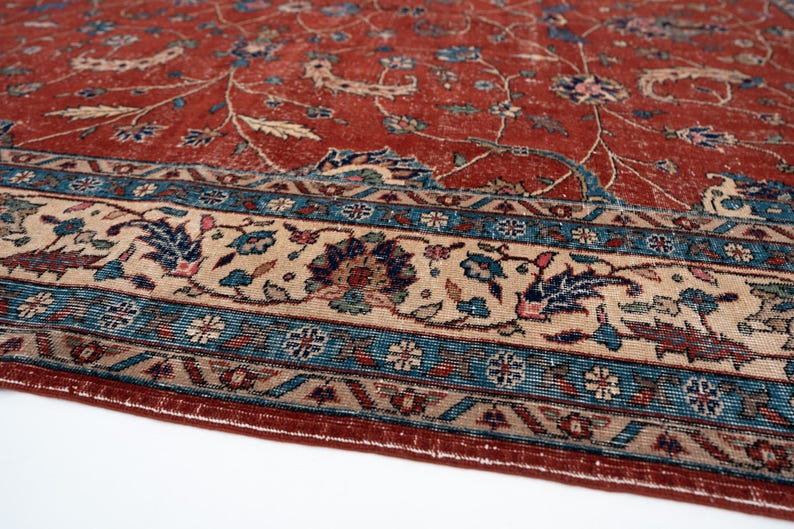 May include: Close-up of a distressed, vintage rug with a rich, reddish-brown field and intricate floral patterns in blue, beige, and other colors. The rug features a detailed border with geometric and floral motifs.