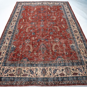 May include: A large, rectangular rug with a central field of faded red, adorned with floral patterns in blue, beige, and hints of green. The rug features a detailed border with similar floral motifs and a contrasting beige background. The overall design suggests a vintage or antique style.