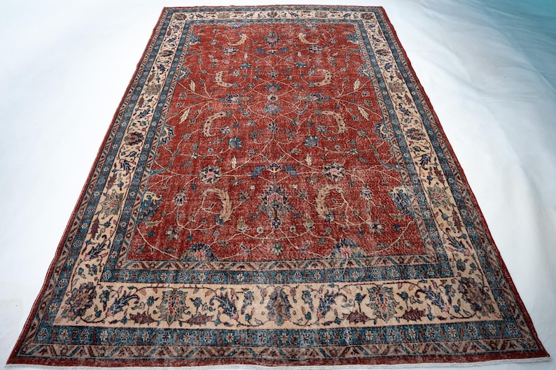 May include: A large, rectangular rug with a central field of faded red, adorned with floral patterns in blue, beige, and hints of green. The rug features a detailed border with similar floral motifs and a contrasting beige background. The overall design suggests a vintage or antique style.
