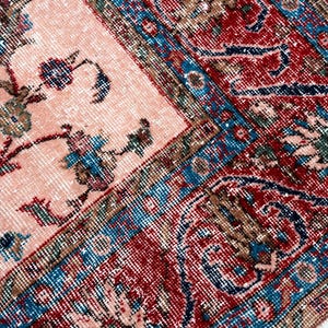 Pu&ograve; includere: Primo piano di un tappeto vintage con un motivo floreale sbiadito nei toni del pesca, rosso, blu e verde. Il tappeto ha un aspetto invecchiato, con un bordo dettagliato e disegni intricati, che mostrano uno stile classico e antico.