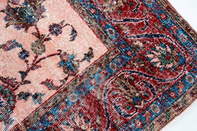Pu&ograve; includere: Primo piano di un tappeto vintage con un motivo floreale sbiadito nei toni del pesca, rosso, blu e verde. Il tappeto ha un aspetto invecchiato, con un bordo dettagliato e disegni intricati, che mostrano uno stile classico e antico.