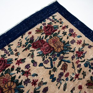 May include: Close-up of a section of a floral rug. The rug features a beige background with a pattern of red, pink, and yellow roses, along with green leaves and small purple flowers. A dark blue border frames the rug.