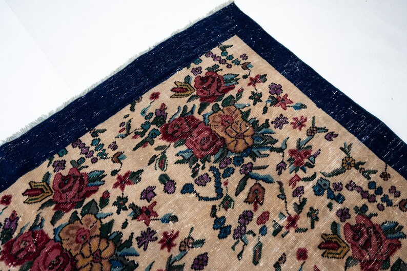 May include: Close-up of a section of a floral rug. The rug features a beige background with a pattern of red, pink, and yellow roses, along with green leaves and small purple flowers. A dark blue border frames the rug.