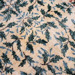 May include: Close-up of a beige rug with a repeating pattern of dark green and light blue leaves and vines. Small clusters of red flowers are visible in the upper right corner. The rug has a distressed, vintage appearance.