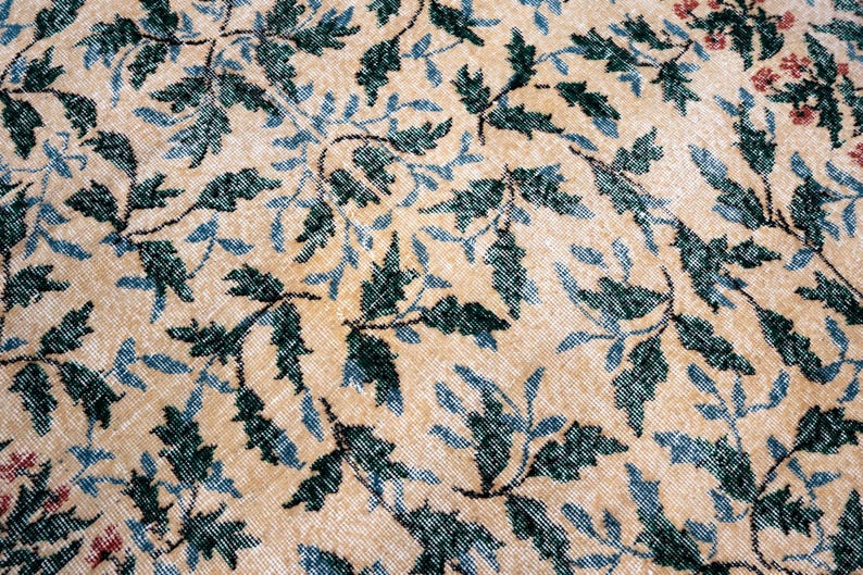 May include: Close-up of a beige rug with a repeating pattern of dark green and light blue leaves and vines. Small clusters of red flowers are visible in the upper right corner. The rug has a distressed, vintage appearance.