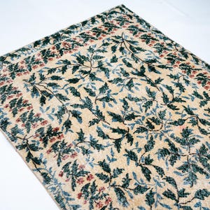 May include: Cream-colored rug with a repeating pattern of green and blue leaves and red floral accents. The rug has a border with a similar floral design. The rug is rectangular and appears to be made of a woven material.