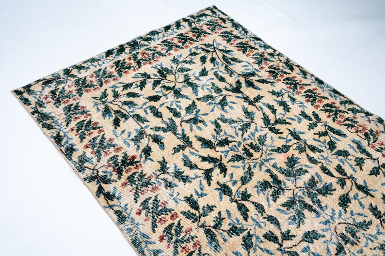 May include: Cream-colored rug with a repeating pattern of green and blue leaves and red floral accents. The rug has a border with a similar floral design. The rug is rectangular and appears to be made of a woven material.
