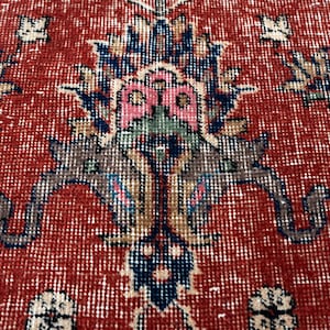 May include: Close-up of a red and white patterned rug with a distressed, vintage look. The rug features a central floral design in shades of blue, pink, and brown. The rug's texture is visible, adding to its aged appearance.