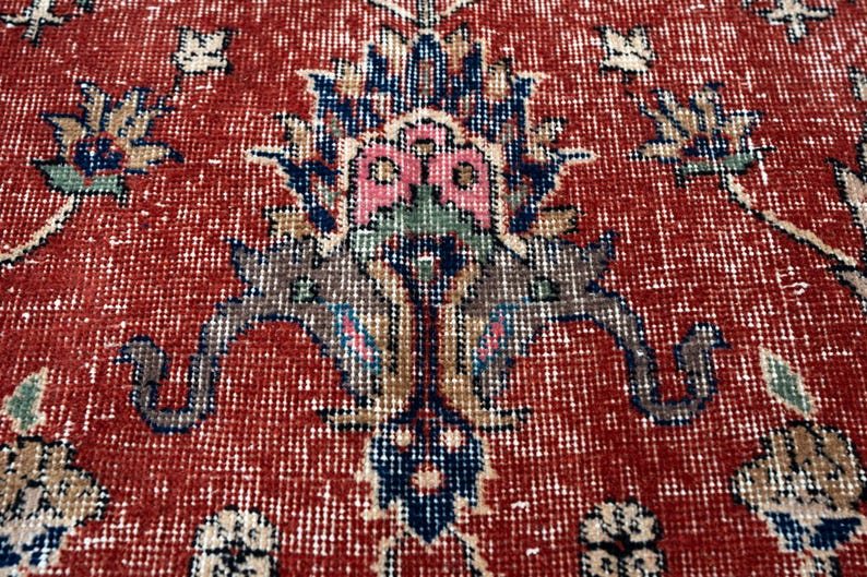May include: Close-up of a red and white patterned rug with a distressed, vintage look. The rug features a central floral design in shades of blue, pink, and brown. The rug's texture is visible, adding to its aged appearance.