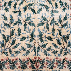 May include: Beige rug with a repeating pattern of teal and blue leaves and red flowers. The floral design is symmetrical, with a border of red flowers. The rug has a vintage, distressed look.