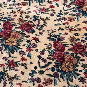 May include: Close-up of a beige floral rug with a detailed pattern. The rug features clusters of red, yellow, and purple flowers with blue and green foliage. The rug's design is intricate and the colors are vibrant.