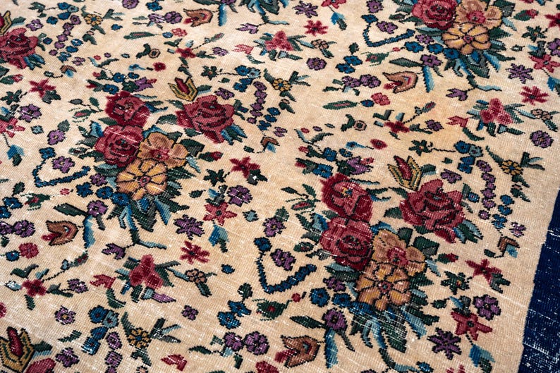 May include: Close-up of a beige floral rug with a detailed pattern. The rug features clusters of red, yellow, and purple flowers with blue and green foliage. The rug's design is intricate and the colors are vibrant.