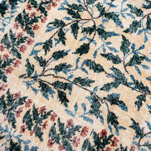 May include: Close-up of a beige rug with a floral pattern. The design features dark green leaves, blue leaves, and small red flowers. The rug has a distressed, vintage look.