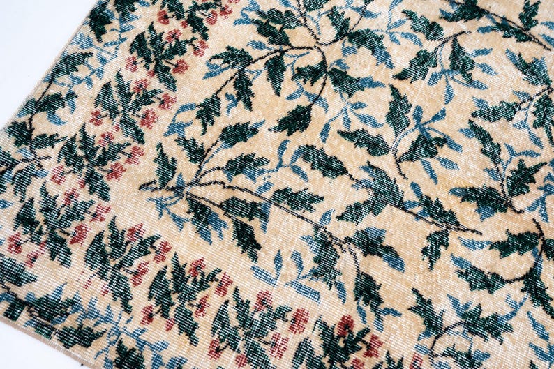 May include: Close-up of a beige rug with a floral pattern. The design features dark green leaves, blue leaves, and small red flowers. The rug has a distressed, vintage look.