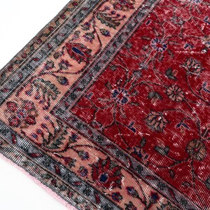 K&ouml;nnte beinhalten: Nahaufnahme eines Abschnitts eines roten, antiken Teppichs mit einem komplizierten Blumenmuster. Der Teppich hat ein verblasstes Aussehen mit einem rosa und grauen Rand. Das Design umfasst blaue und braune Akzente und einen Fransenrand.