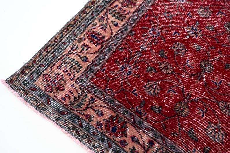 K&ouml;nnte beinhalten: Nahaufnahme eines Abschnitts eines roten, antiken Teppichs mit einem komplizierten Blumenmuster. Der Teppich hat ein verblasstes Aussehen mit einem rosa und grauen Rand. Das Design umfasst blaue und braune Akzente und einen Fransenrand.