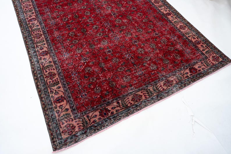 K&ouml;nnte beinhalten: Ein rechteckiger Teppich im Vintage-Stil mit einem tiefroten Feld und einem floralen Muster. Der Rand weist ein erg&auml;nzendes Design in Rosa-, Blau- und Gr&uuml;nt&ouml;nen auf. Der Teppich liegt auf einer wei&szlig;en Oberfl&auml;che.