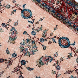 Pu&ograve; includere: Primo piano di un tappeto vintage dall'aspetto sbiadito e usurato. Il tappeto presenta uno sfondo color pesca chiaro con un motivo floreale in verde acqua, blu e rosso tenue. Il bordo &egrave; rosso pi&ugrave; scuro con disegni geometrici.