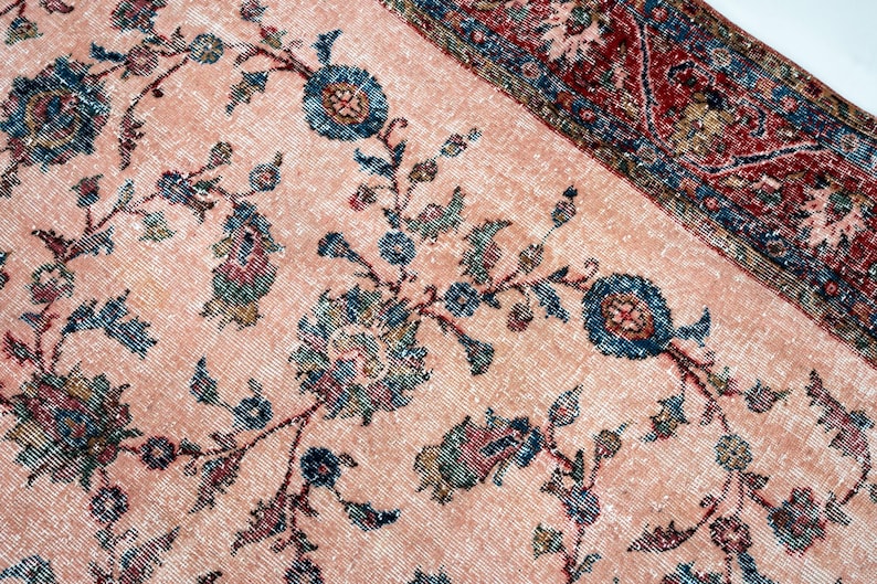Pu&ograve; includere: Primo piano di un tappeto vintage dall'aspetto sbiadito e usurato. Il tappeto presenta uno sfondo color pesca chiaro con un motivo floreale in verde acqua, blu e rosso tenue. Il bordo &egrave; rosso pi&ugrave; scuro con disegni geometrici.