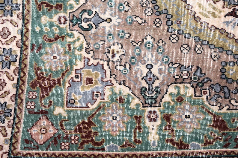 May include: A woven rug featuring a complex geometric and floral pattern in shades of teal, taupe, cream, and dark brown. The design includes intricate borders and central motifs with repeating symmetrical shapes and detailed botanical elements.