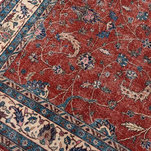 May include: A close-up of a vintage rug with a distressed, faded appearance. The rug features a deep red central field with floral patterns in blue, cream, and pink. A cream border with blue and floral designs frames the rug.