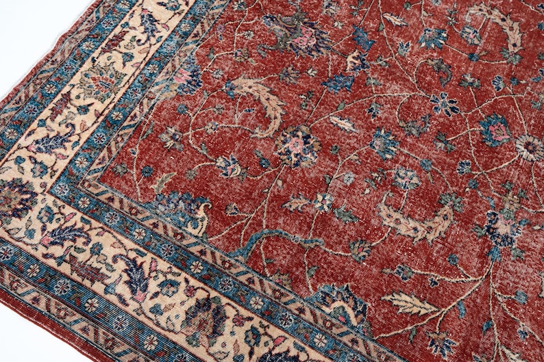 May include: A close-up of a vintage rug with a distressed, faded appearance. The rug features a deep red central field with floral patterns in blue, cream, and pink. A cream border with blue and floral designs frames the rug.