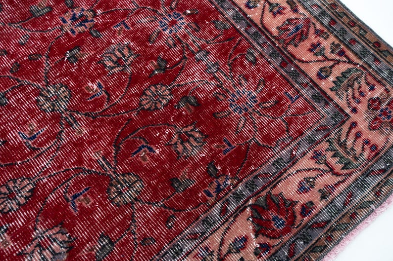 K&ouml;nnte beinhalten: Nahaufnahme eines abgenutzten roten Teppichs mit floralen Mustern. Der Teppich hat ein rotes Mittelfeld mit verblassten Blumenmotiven in Blau, Grau und Creme. Ein Rand in Rosa- und Graut&ouml;nen umrahmt das Design und erzeugt eine Vintage-&Auml;sthetik.