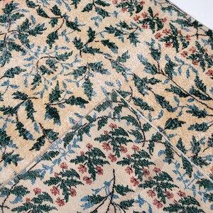 May include: Beige rug with a repeating pattern of green leaves, blue accents, and small red flowers. The rug has a distressed, vintage look, with a folded corner revealing the underside.