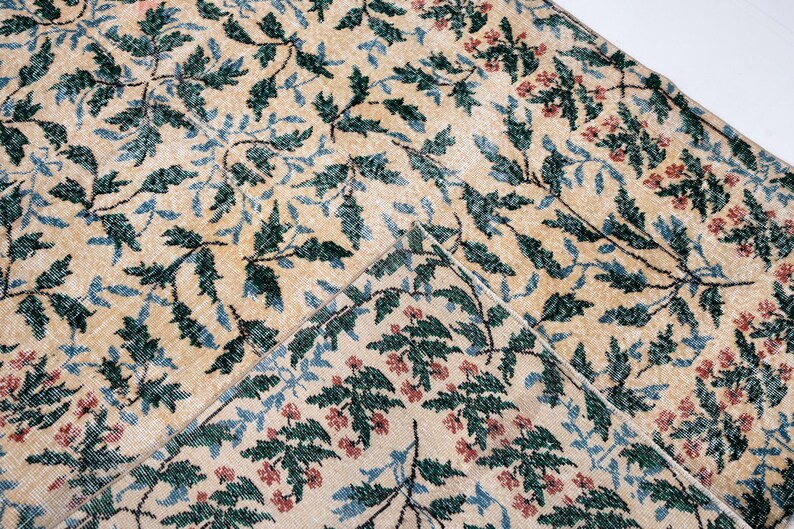 May include: Beige rug with a repeating pattern of green leaves, blue accents, and small red flowers. The rug has a distressed, vintage look, with a folded corner revealing the underside.