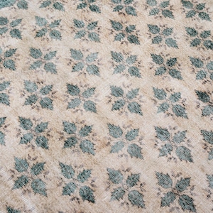 May include: A rectangular area rug featuring a repeating pattern of dark teal, four-leaf floral motifs arranged in a grid across a light beige, textured background. The rug is framed by a solid dark teal border along the visible top edge.