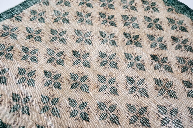 May include: A rectangular area rug featuring a repeating pattern of dark teal, four-leaf floral motifs arranged in a grid across a light beige, textured background. The rug is framed by a solid dark teal border along the visible top edge.