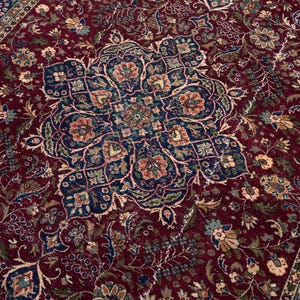 May include: A detailed close-up of a Persian-style rug. The rug features a deep burgundy field with an intricate central medallion in navy blue, accented by floral and geometric patterns in beige, green, and orange. The border is navy blue with floral motifs.