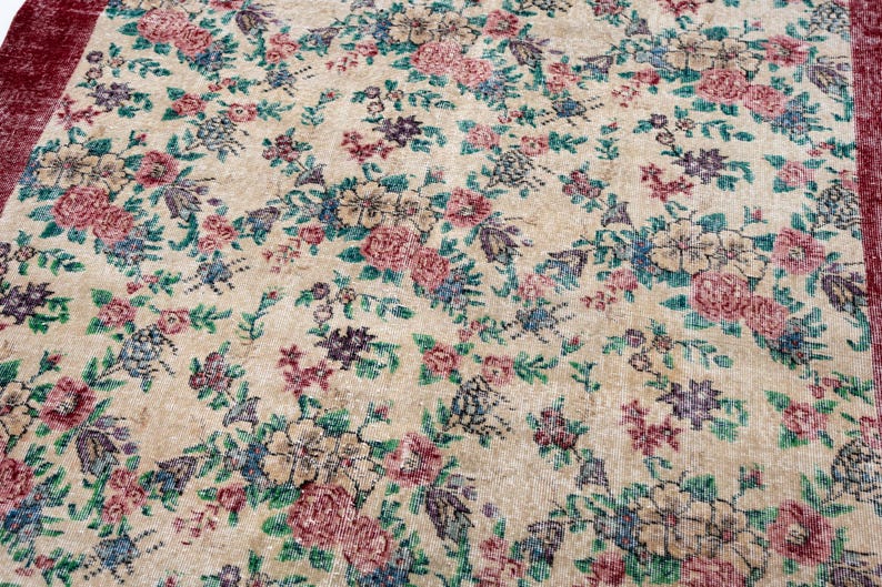 Pode incluir: Um tapete vintage com fundo bege e padr&atilde;o floral. O tapete apresenta flores rosa, azuis e roxas com folhas verdes. A borda &eacute; de cor bord&ocirc; escuro. Este tapete &eacute; adequado para salas de estar e quartos.