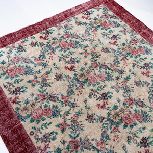 Pode incluir: Um tapete retangular com fundo creme e um padr&atilde;o floral em tons de rosa, azul e verde. O tapete tem uma borda vermelha escura. O tapete &eacute; provavelmente feito de um material tecido.