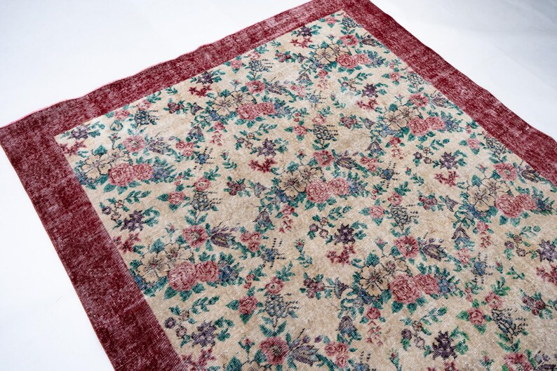 Pode incluir: Um tapete retangular com fundo creme e um padr&atilde;o floral em tons de rosa, azul e verde. O tapete tem uma borda vermelha escura. O tapete &eacute; provavelmente feito de um material tecido.