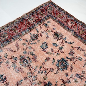 Pu&ograve; includere: Un angolo di un tappeto vintage con sfondo color pesca sbiadito e intricati motivi floreali in turchese, blu e bordeaux. Il bordo presenta un design dettagliato nei toni del rosso e del blu, mostrando un'estetica classica e invecchiata.