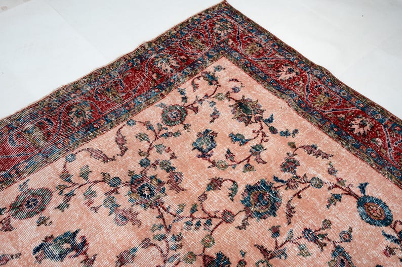 Pu&ograve; includere: Un angolo di un tappeto vintage con sfondo color pesca sbiadito e intricati motivi floreali in turchese, blu e bordeaux. Il bordo presenta un design dettagliato nei toni del rosso e del blu, mostrando un'estetica classica e invecchiata.