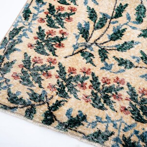 May include: Close-up of a section of a beige rug with a floral pattern. The design features green leaves, red flowers, and blue accents. The rug has a distressed, vintage appearance, with a faded look.