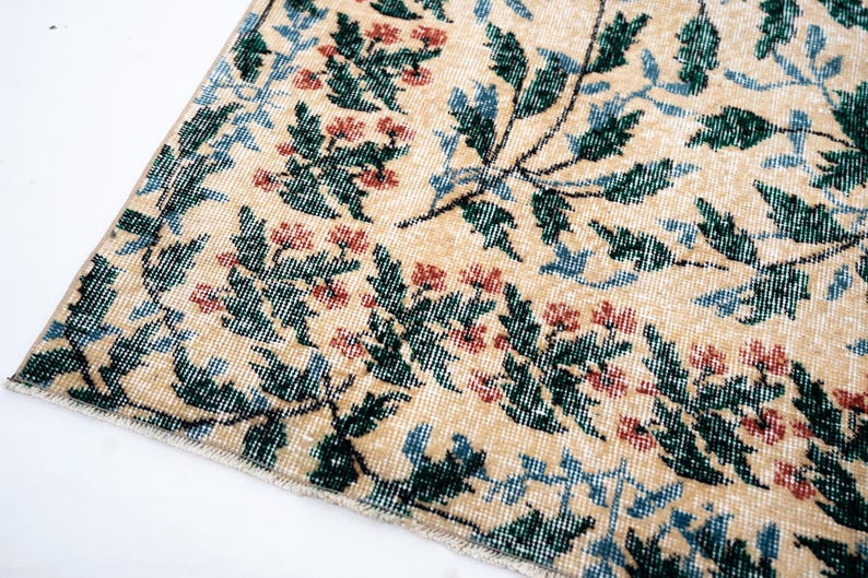 May include: Close-up of a section of a beige rug with a floral pattern. The design features green leaves, red flowers, and blue accents. The rug has a distressed, vintage appearance, with a faded look.