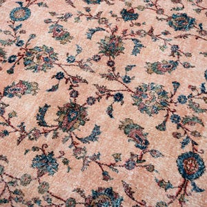 Pu&ograve; includere: Un tappeto vintage dall'aspetto sbiadito e usurato. Il tappeto presenta un campo centrale color pesca adornato da un motivo floreale in verde acqua, blu e rosso tenue. Il bordo &egrave; rosso scuro con disegni intricati.