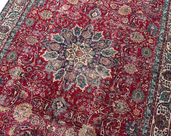 May include: A rectangular area rug featuring a deep red background with a central dark blue and cream floral medallion. The surface is covered in intricate scrolling vine and leaf patterns in shades of teal, navy, and beige, framed by a wide border.