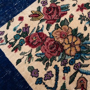 May include: Close-up of a section of a floral rug with a navy blue border. The rug features a cream-colored background with a pattern of red, yellow, and purple flowers and green leaves. The rug has a vintage, handmade appearance.