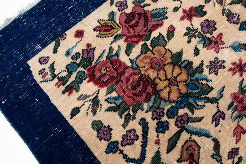 May include: Close-up of a section of a floral rug with a navy blue border. The rug features a cream-colored background with a pattern of red, yellow, and purple flowers and green leaves. The rug has a vintage, handmade appearance.