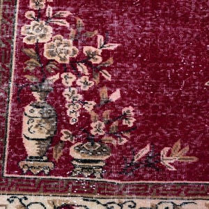 May include: Close-up of a red and beige floral patterned rug. The rug features a central red field with floral and vase motifs in beige and brown. The border is beige with a repeating pattern. The rug has a distressed, vintage appearance.