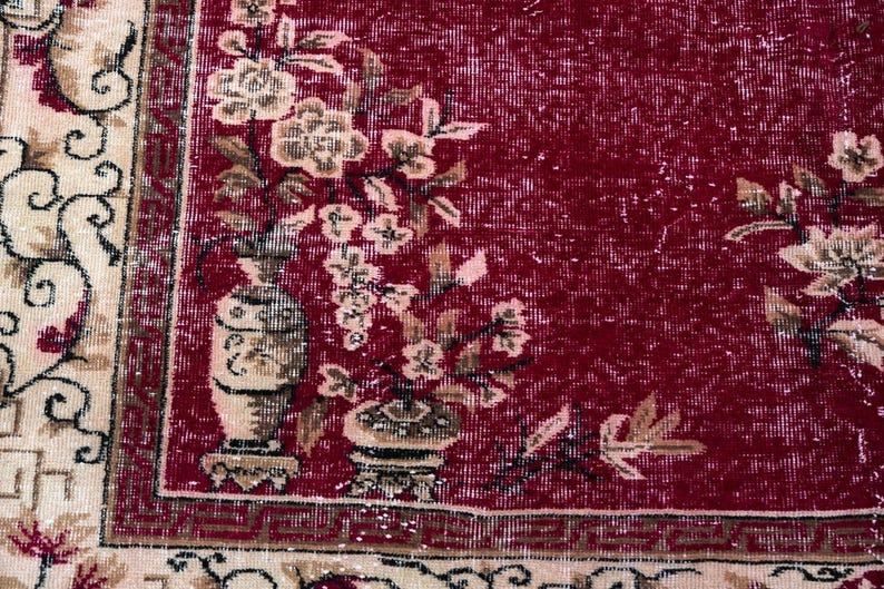 May include: Close-up of a red and beige floral patterned rug. The rug features a central red field with floral and vase motifs in beige and brown. The border is beige with a repeating pattern. The rug has a distressed, vintage appearance.