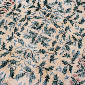 May include: Close-up of a beige rug with a repeating pattern of green and blue leaves and small red flowers. The rug has a distressed, vintage look, with a soft, faded color palette. The rug is likely made of wool or a similar natural fiber.