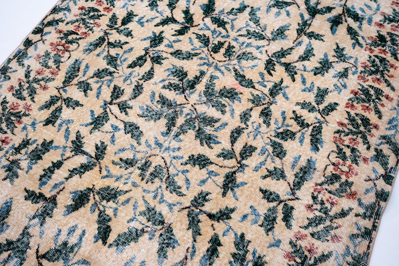 May include: Close-up of a beige rug with a repeating pattern of green and blue leaves and small red flowers. The rug has a distressed, vintage look, with a soft, faded color palette. The rug is likely made of wool or a similar natural fiber.